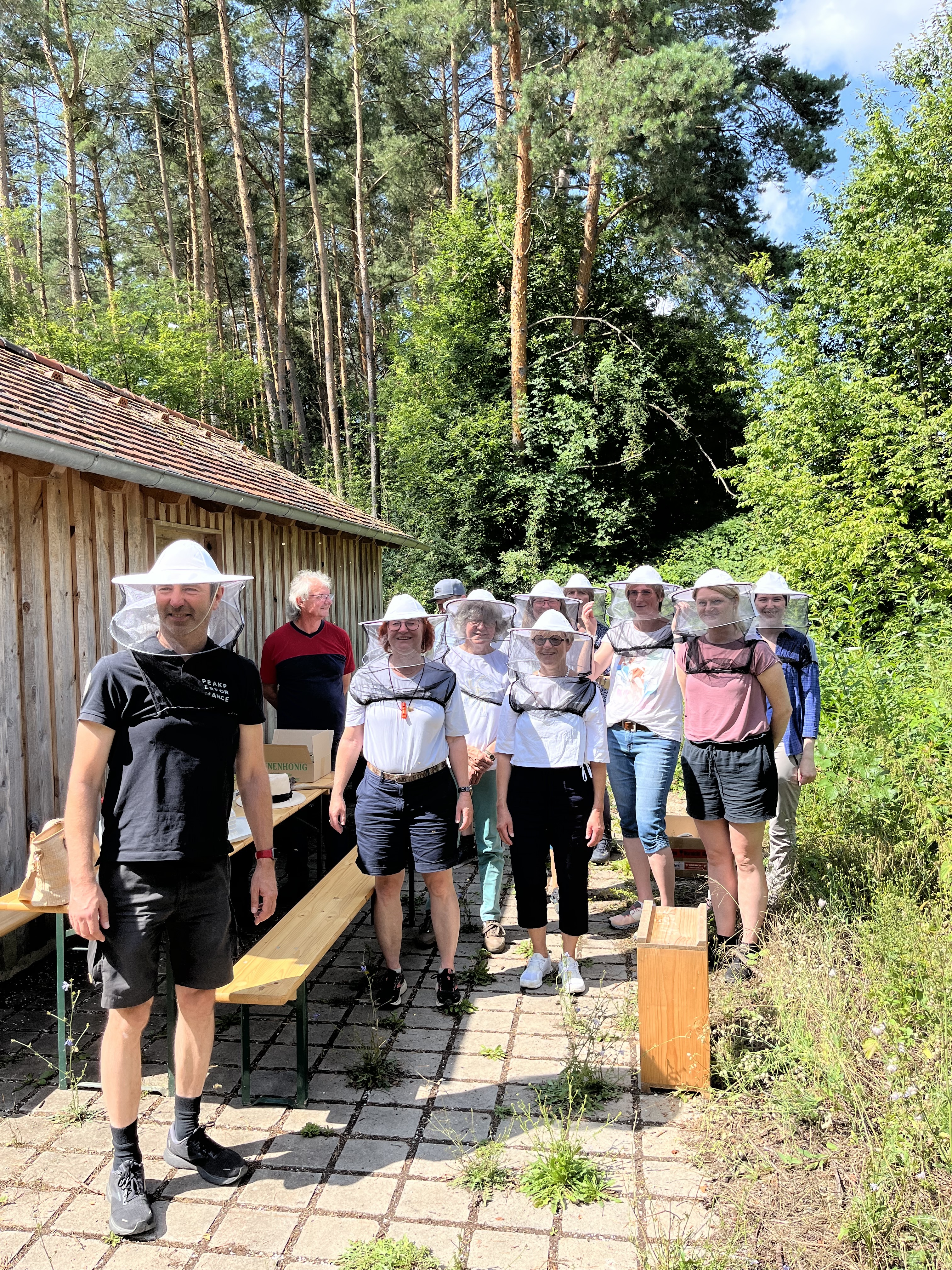 Personen mit Bienenschutzhauben stehen neben einer Hütte umgeben von Bäumen, Foto cm