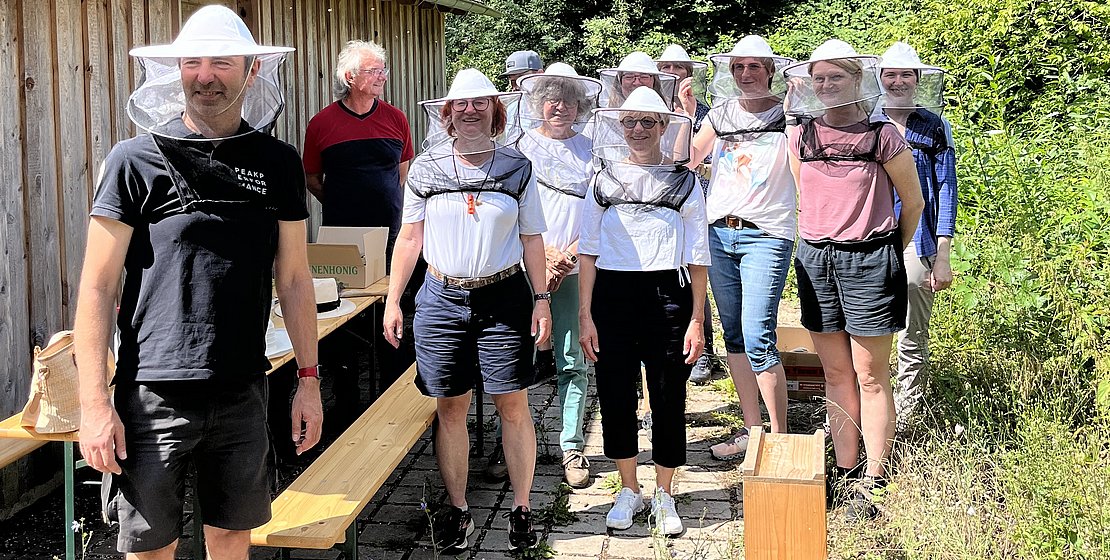 Personen mit Bienenschutzhauben stehen neben einer Hütte umgeben von Bäumen, Foto cm