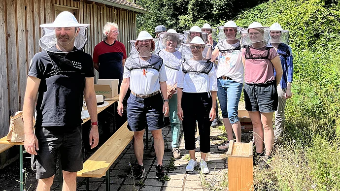 Personen mit Bienenschutzhauben stehen neben einer Hütte umgeben von Bäumen, Foto cm